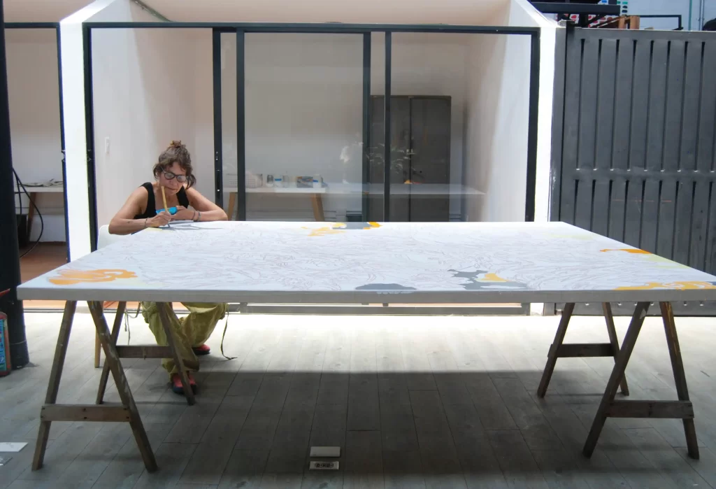 Artista concentrada pintando una gran superficie blanca con líneas y figuras en tonos amarillos y grises, en un estudio bien iluminado y moderno.