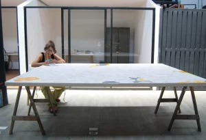 Artista concentrada pintando una gran superficie blanca con líneas y figuras en tonos amarillos y grises, en un estudio bien iluminado y moderno.