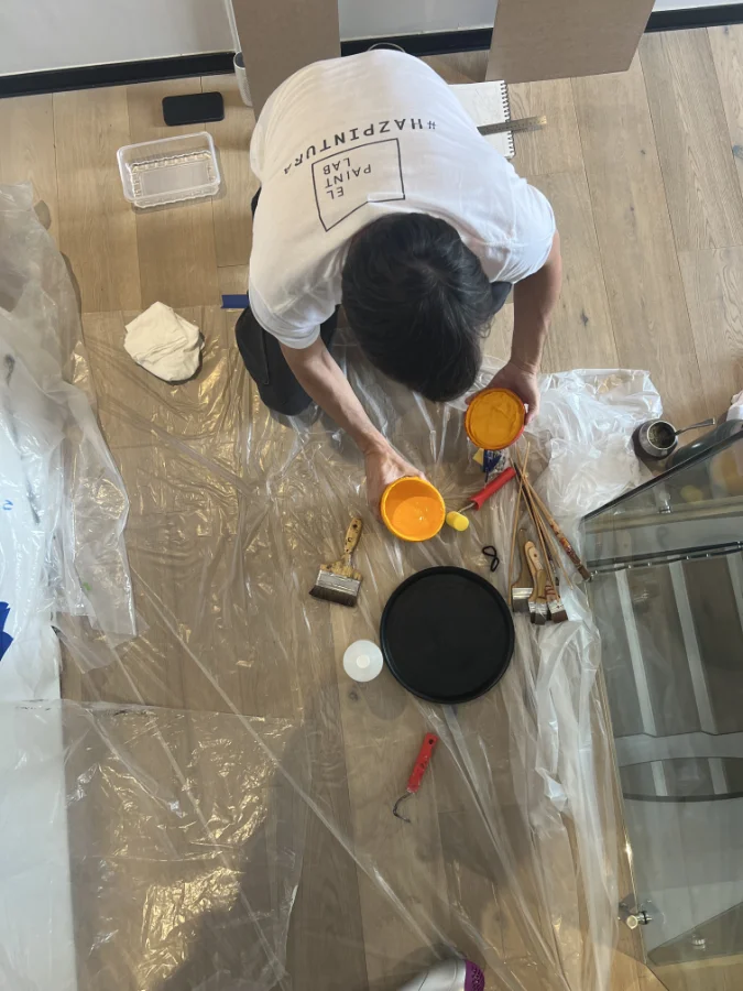 Artista visto desde arriba preparando pintura amarilla en el suelo, rodeado de brochas, herramientas y pigmentos del Paintlab para pintar.
