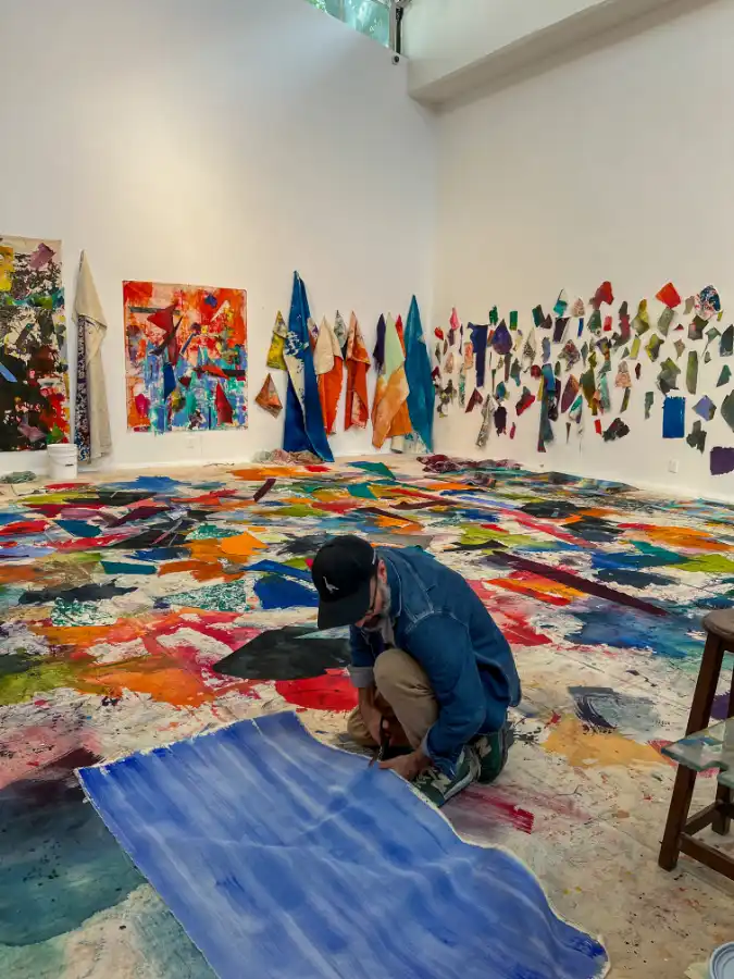 Artista patrocinado por El Paint Lab trabajando en una pieza sobre tela pintada, rodeado de retazos coloridos en el suelo y paredes de un estudio artístico.