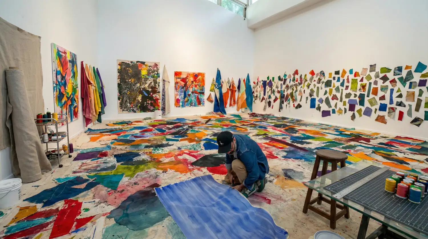 Artista patrocinado por El Paint Lab trabajando en una pieza sobre tela pintada, rodeado de retazos coloridos en el suelo y paredes de un estudio artístico.
